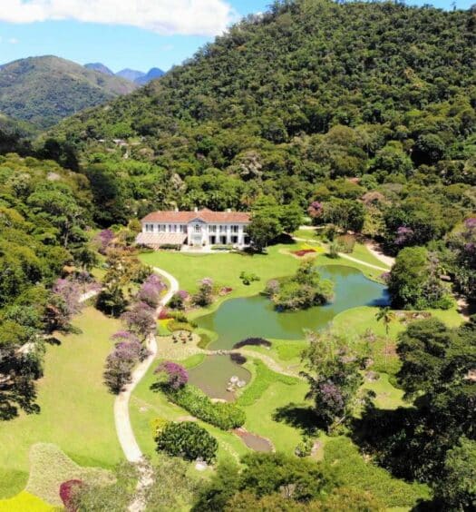 Vista aérea da propriedade Casa Marambaia Hotel, com uma construção estilo colonial ao centro, cercada por montanhas e árvores, com um extenso lago do lado direito, para representar hotéis em Petrópolis