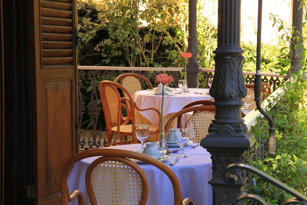 Varanda com mesas para refeições do Casablanca Koeler com mesas redondas e cadeiras de madeira, ao redor, há muito verde e árvores