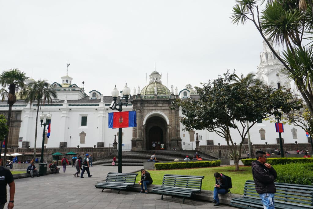 Praça em Quito, no Equador, com pessoas andando e outras sentadas. Há uma catedral ao fundo com vários degraus na frente, a bandeira da cidade pendurada em um poste de luz e um jardim ao lado esquerdo com grama baixa e árvores.
