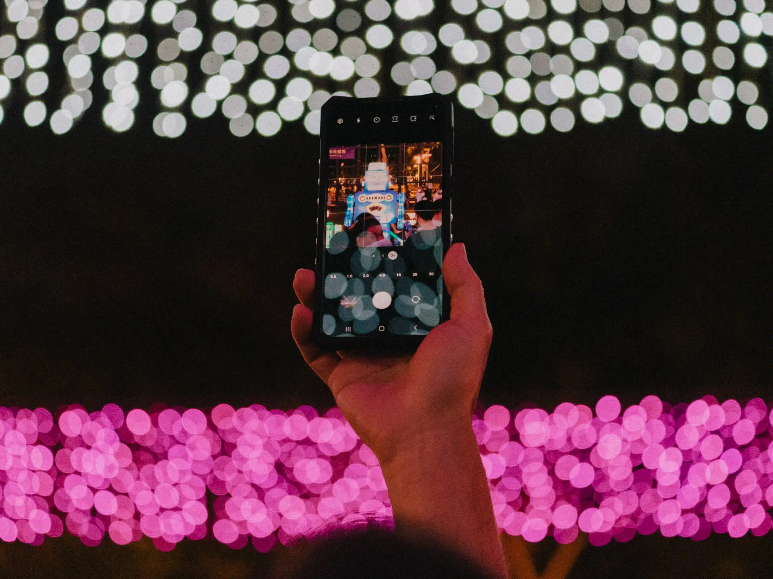 Uma mão segurando o celular em Hing Fat Street, Hong Kong para tirar foto durante a noite com várias luzes. Representa chip celular Hong Kong.