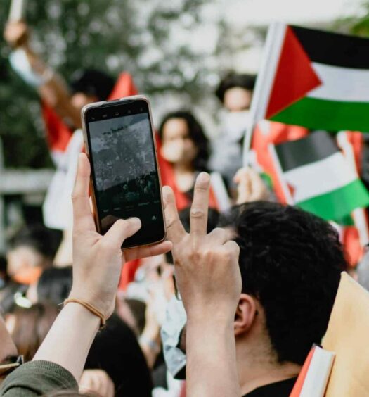 Celular na mão de um homem, com pessoas em volta com a bandeira da Palestina. Imagem para ilustrar o post chip de celular Palestina