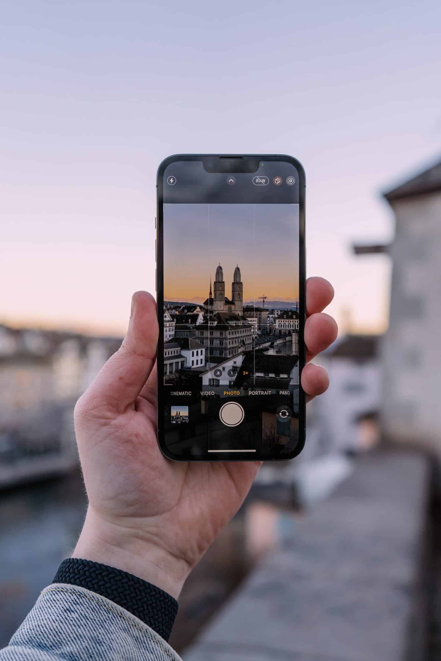 Uma mão segurando um celular tirando foto do centro histórico de Zurique, com prédios antigos e pôr do sol ao fundo. Imagem para ilustrar o post chip de celular Zurique