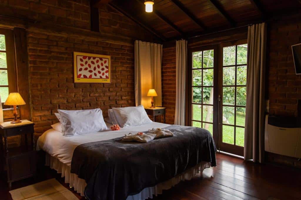 Quarto na Pousada das Araras com chão de madeira, uma cama de casal, paredes de tijolinhos e uma porta de vidro que leva para o jardim
