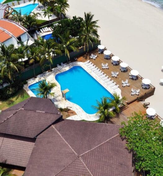 Vista aérea da Coconut's Maresias Hotel com a piscina e o deck dela bem pertinho da areia da praia, onde há algumas cadeiras e guarda-sóis, para representar pousadas pé na areia em Maresias
