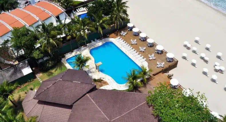 Vista aérea da Coconut's Maresias Hotel com a piscina e o deck dela bem pertinho da areia da praia, onde há algumas cadeiras e guarda-sóis, para representar pousadas pé na areia em Maresias