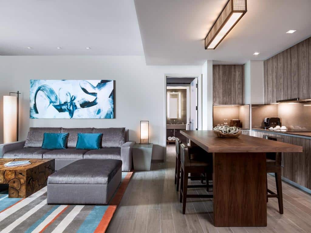Sala de estar e cozinha comacta do East Miami com sofá cinza com almofadas azul turquesa no lado esquerdo com mesa de madeira ao centro. Do lado direito mesa de três lugares com armários ao fundo. Representa hotéis com cozinha em Miami.