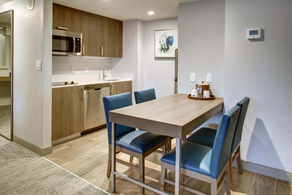 Cozinha do Hampton Inn & Suites Miami Airport South/Blue Lagoon com mesa quadrada de quatro cadeiras azuis, ao fundo armário de madeira com fogão, pia e micro-ondas. Representa hotéis com cozinha em Miami.