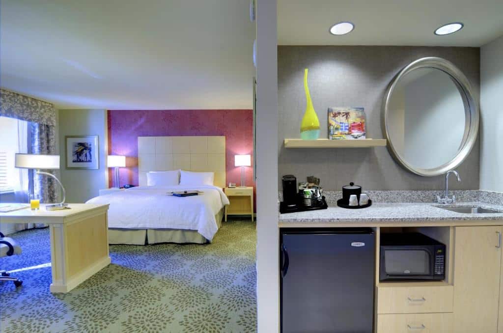 Quarto do Hampton Inn & Suites by Hilton Miami Downtown/Brickell com cama de casal, duas cômodas com luminárias, mesa de trabalho a frente do lado direito cozinha compacta com pia de mármore, micro-ondas e frigobar.