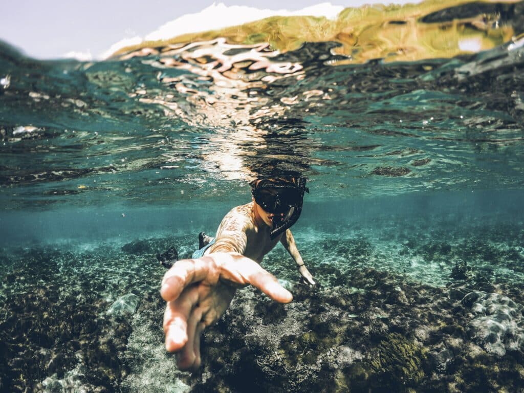 homem branco debaixo d'água na ilha de Lembongan, Indonesia. A foto mostra um homem nadando no centro da imagem, olhando para a câmera e esticando a mão para alcançar o aparelho fotográfico. Na parte superior da foto é possível ver um pouco da terra fora d'água
