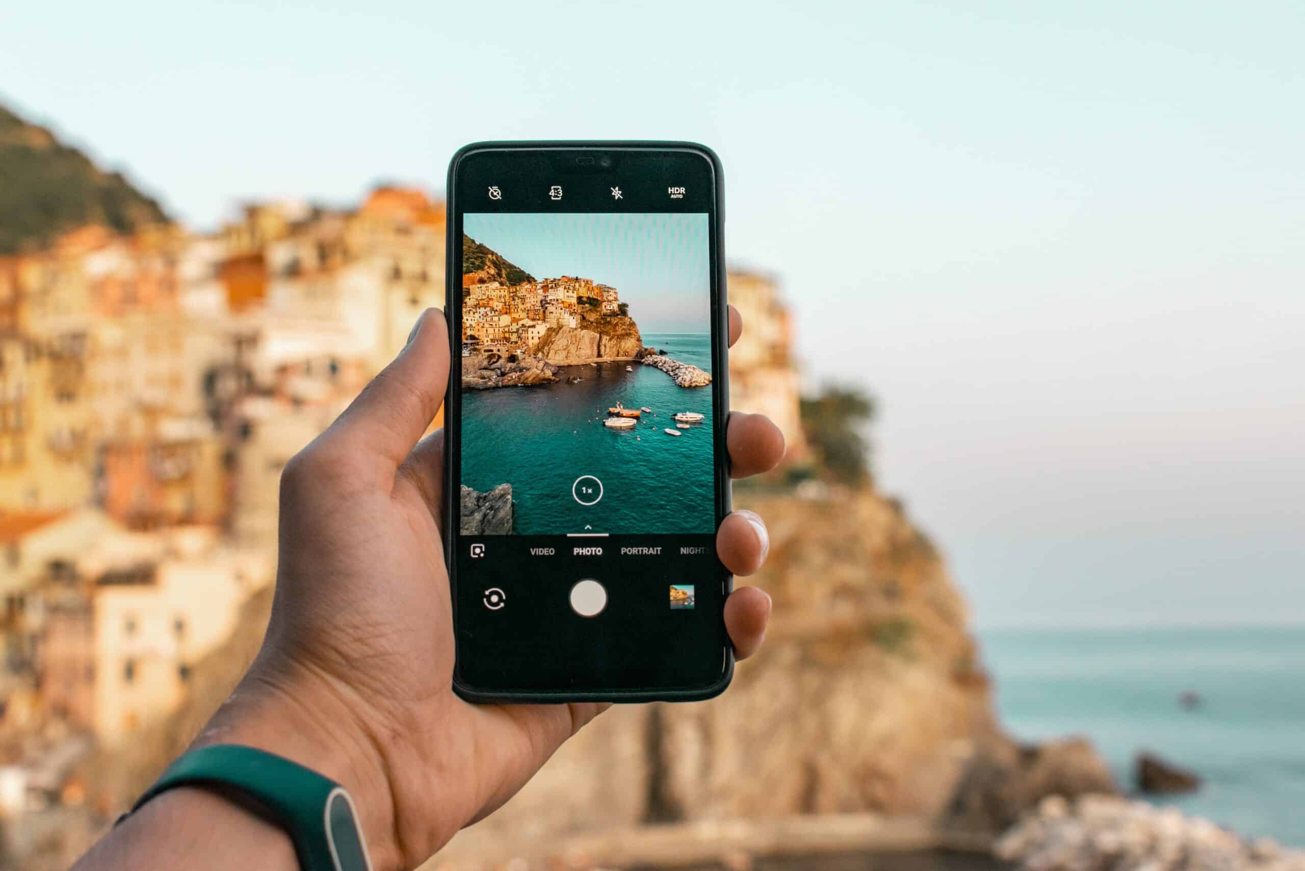 mão masculina segurando um aparelho iphone para tirar a foto de uma encosta italiana, mostrando as casas e os barcos na água azul, ao fim de tarde com a luz do sol refletindo na vila