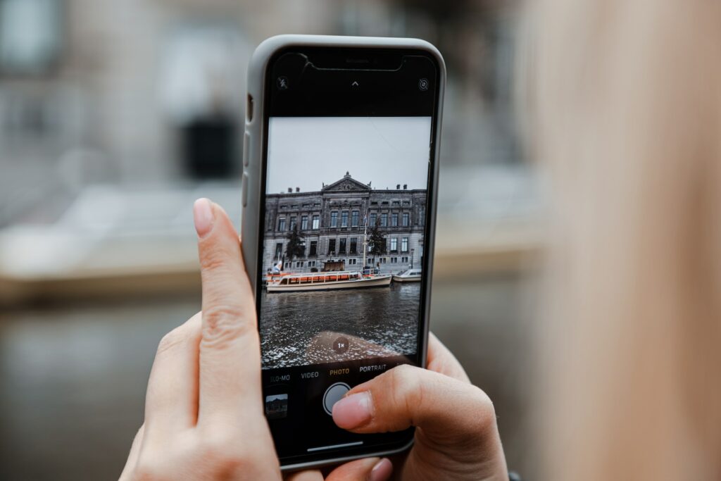 Uma mão branca segurando um celular enquanto fotografa um prédio antigo de concreto que está nas margens de um dos canais de Amsterdam