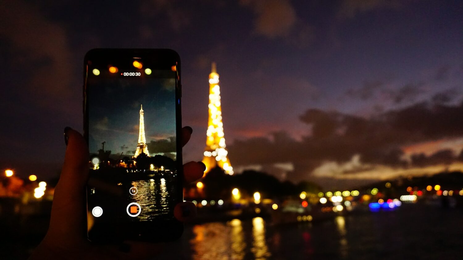 Uma torre alta e iluminada por luzes amarelas, na frente da construção há um rio, e do outro lado deste rio há uma pessoa segurando um celular enquanto fotografa a atração turística, para representar chip celular Paris