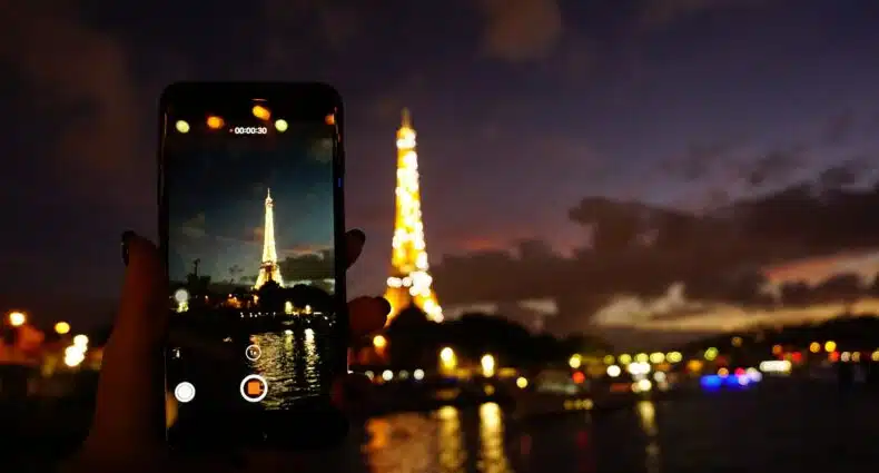 Uma torre alta e iluminada por luzes amarelas, na frente da construção há um rio, e do outro lado deste rio há uma pessoa segurando um celular enquanto fotografa a atração turística, para representar chip celular Paris