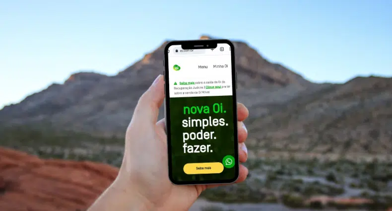 mão de uma pessoa branca segurando um celular com o print do chip Oi em tons de verde com montanhas e céu azul ao fundo meio desfocado