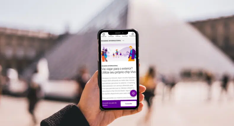 pessoa segurando um celular com a página inicial da Vivo com o Museu do Louvre, em Paris, França atrás desfocado, que é um prédio em forma de pirâmide de vidro, para ilustrar o post de chip Vivo no exterior