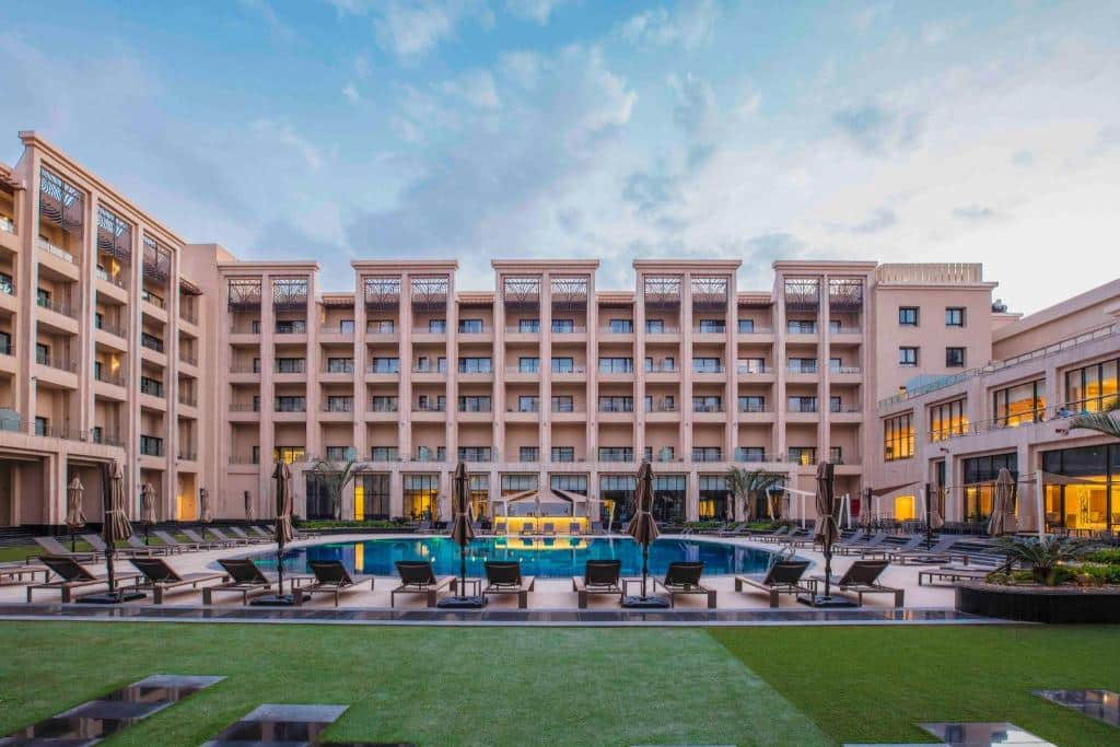 Frente do Triumph Luxury Hotel com piscina a frente com cadeiras em volta e ao fundo a hospedagem. Representa Hotéis no Cairo.