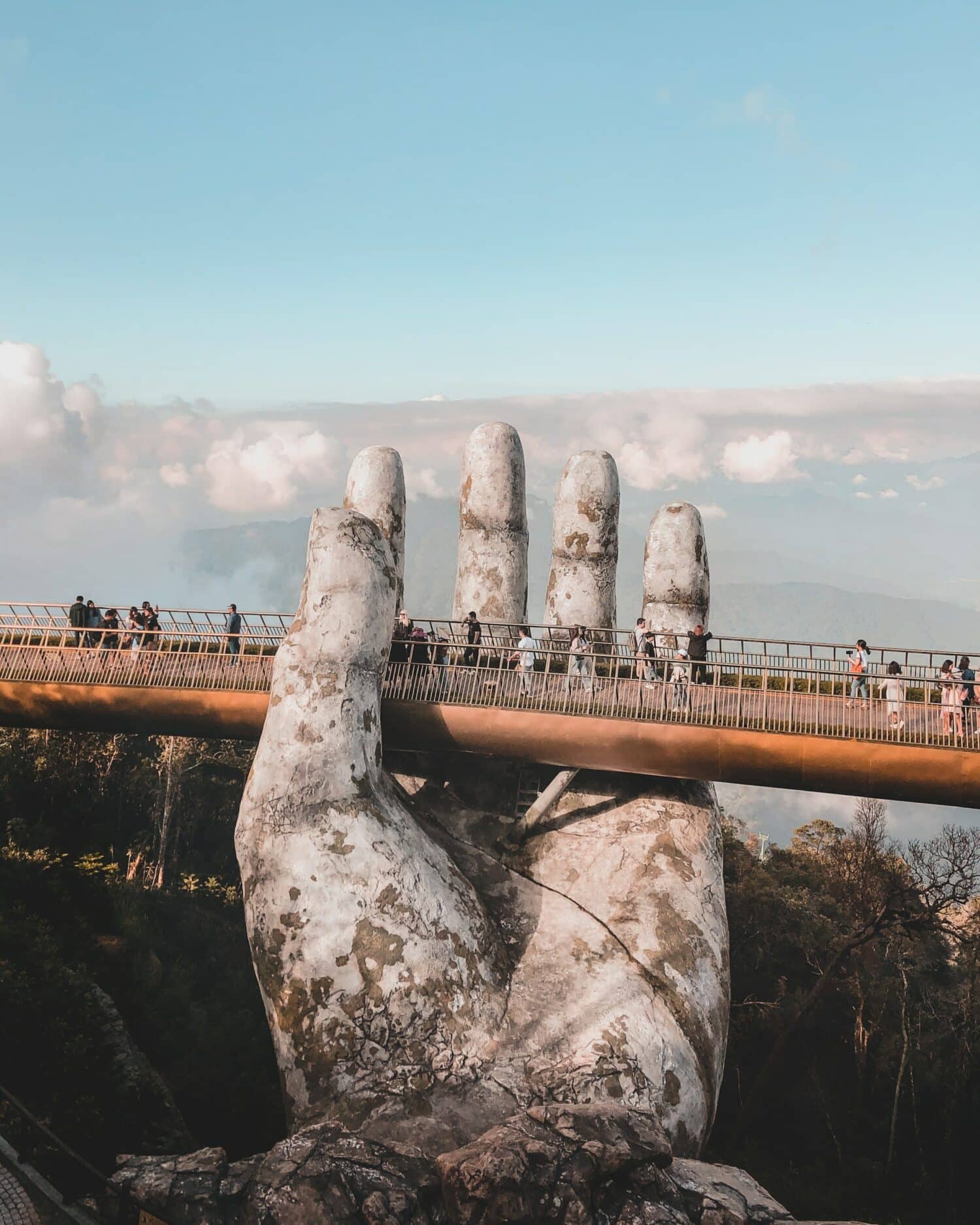 vista de uma ponte, a Golden Bridge, em Danang, com várias pessoas tirando fotos e andando por ela, com vegetação abaixo e um céu azul com nuvens acima, a ponte é "segurada" por uma mão gigantesca de pedra branca, para ilustrar o post de chip celular Vietnã