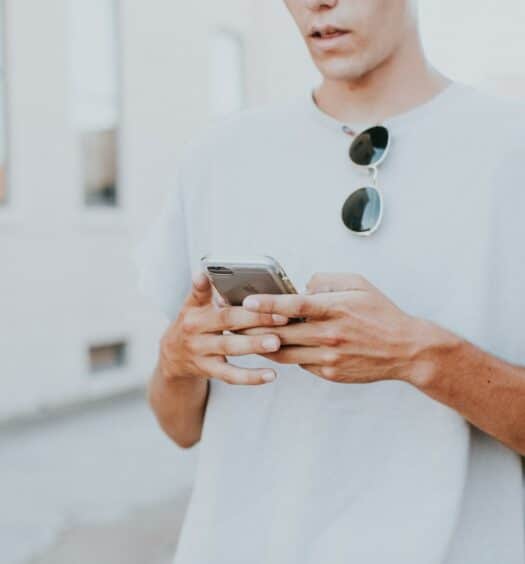 Homem branco mexendo no celular de frente para a câmera. Ele veste uma camiseta branca e tem um óculos de sol pendurado na gola da blusa. - Foto: Hannah Busing via Unsplash