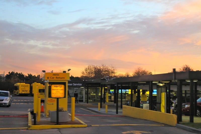 Céu alaranjado em destaque no estacionamento da Hertz Rent a Car do Aeroporto de LaGuardia, em Nova York, nos Estados Unidos. No primeiro plano aparece uma guarida da empresa, um carro vindo em direção a ela, e ao fundo há ônibus da marca Hertz.