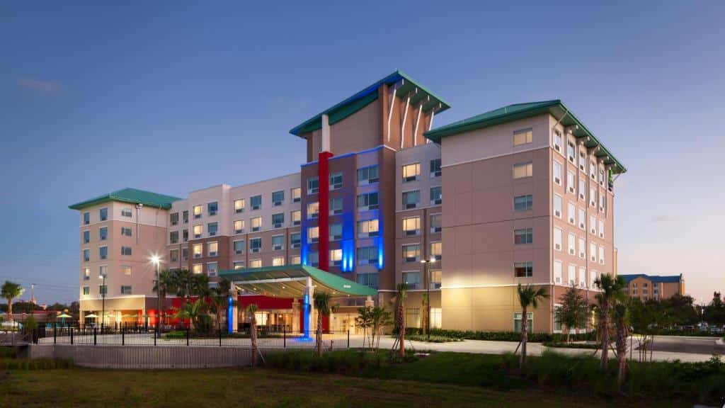 Prédio do Holiday Inn Express & Suites - Orlando At Seaworld com cinco andares, telhado verde e algumas árvores na entrada do hall