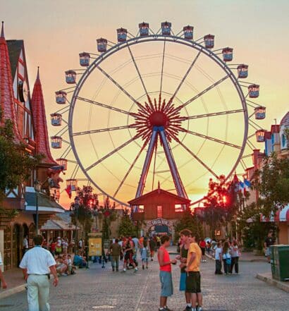 Uma roda gigante colorida ao fundo, na frente há algumas pessoas andando pelo parque, para representar hotéis perto do Hopi Hari