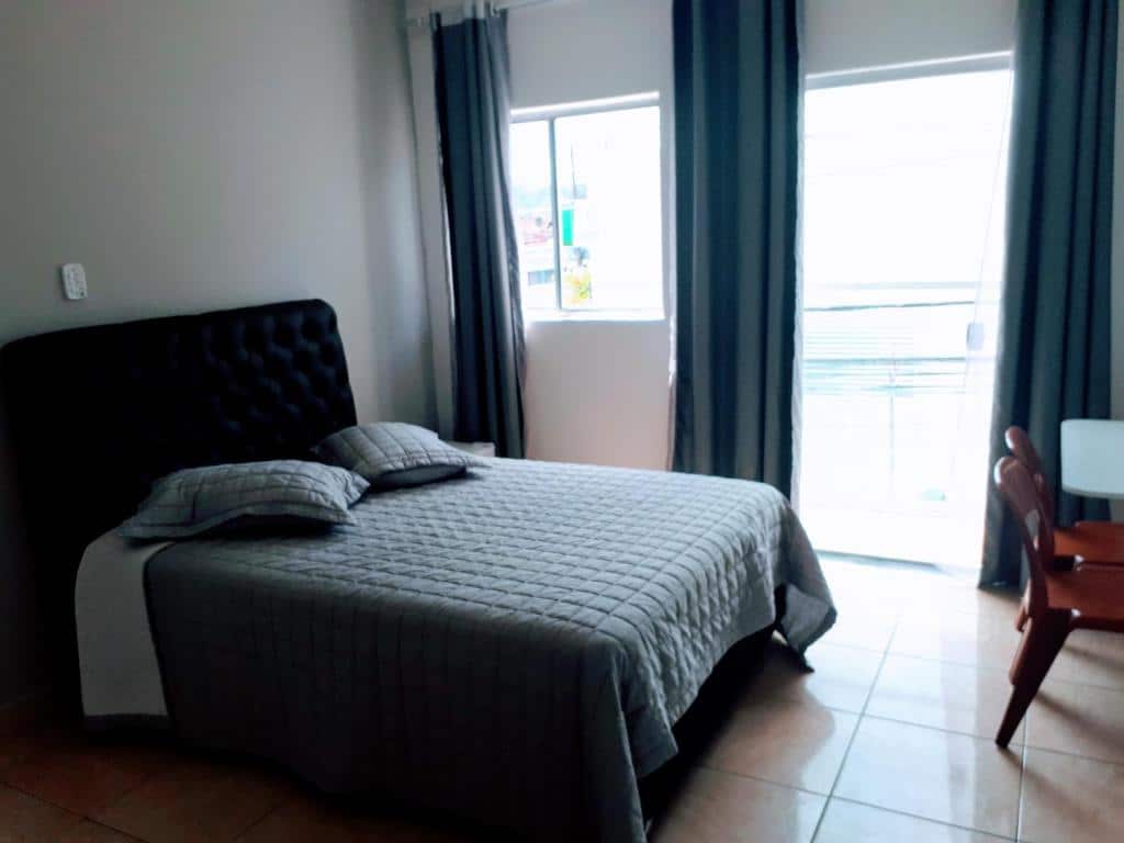 Quarto com cama de casal da Hospedaria São Benedito, com dois travesseiros em cima, duas cadeiras de madeira e uma janela e porta de vidro no lado esquerdo, com cortinas verde escuro
