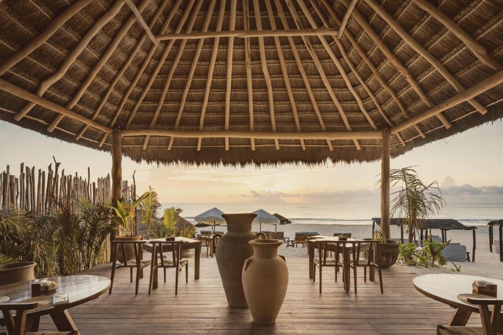 Deck de madeira à beira-mar do Hotel Ma'xanab Tulum, com mesas e cadeiras, vasos artesanais decorando o espaço, além de espreguiçadeiras e guarda-sóis na areia da praia, durante um fim de tarde com um pequeno pôr do sol