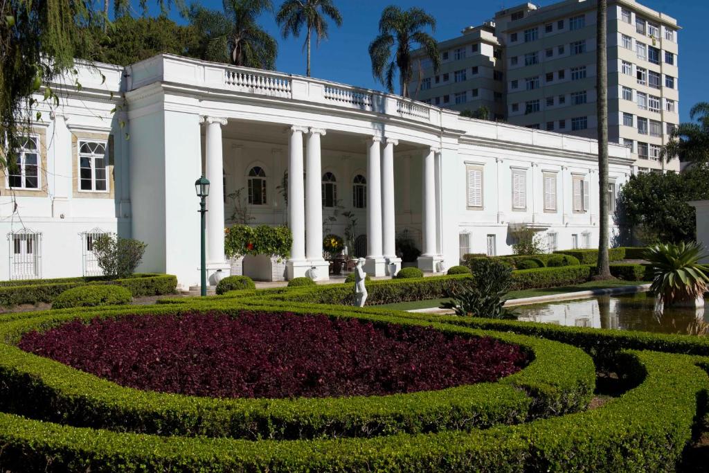 Fachada do Hotel Solar do Império com um amplo jardim na frente, um lago, algumas pequenas esculturas e árvores, para representar hotéis em Petrópolis