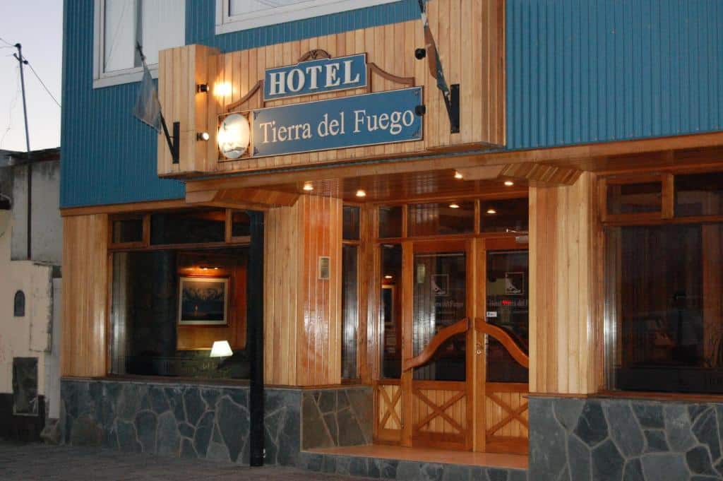 frente do Hotel Tierra del Fuego com o nome pregado acima da porta, em madeira. É possível enxergar as cores azuis na parede externa e vários elementos em madeira rústica