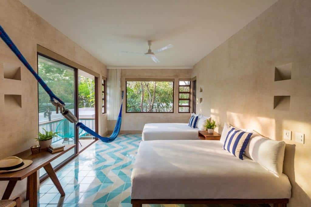 Quarto do Tiki Tiki Tulum, de 30 m², com duas camas de casal, rede azul pendurada de ponta à ponta na frente das camas, mesa com vaso de flor e chapeu em cima, e uma varanda com cadeira azul e vista para a natureza