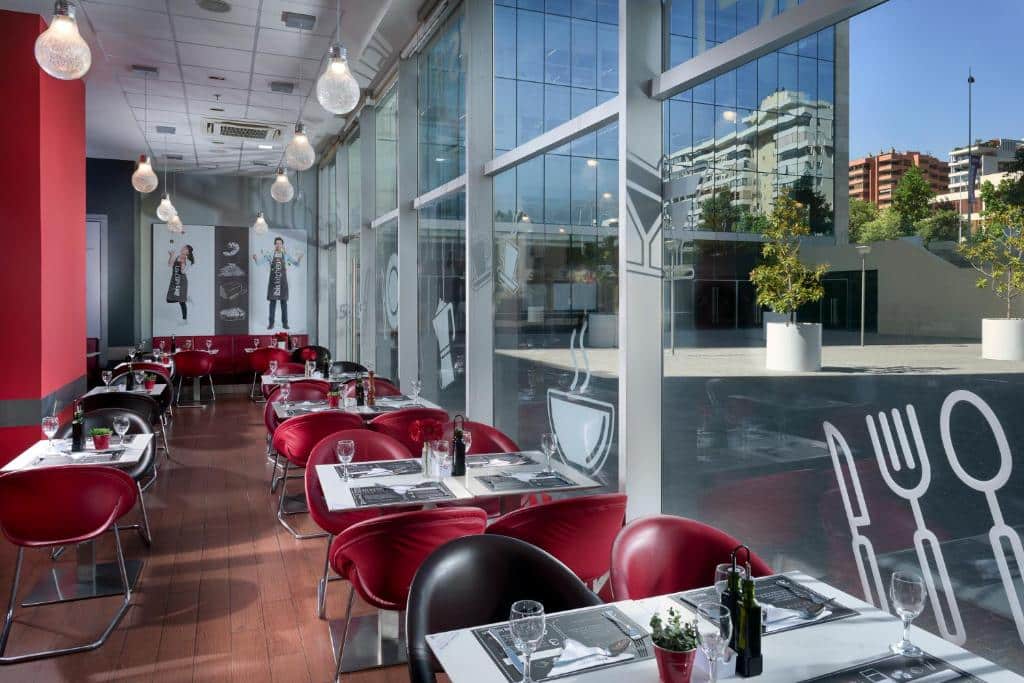 Área de refeições do ibis Santiago Las Condes Manquehue com mesas de metal, cadeiras vermelhas e pretas, ao lado, há uma parede envidraçada com vista para a entrada do hotel, para representar hotéis Ibis em Santiago do Chile