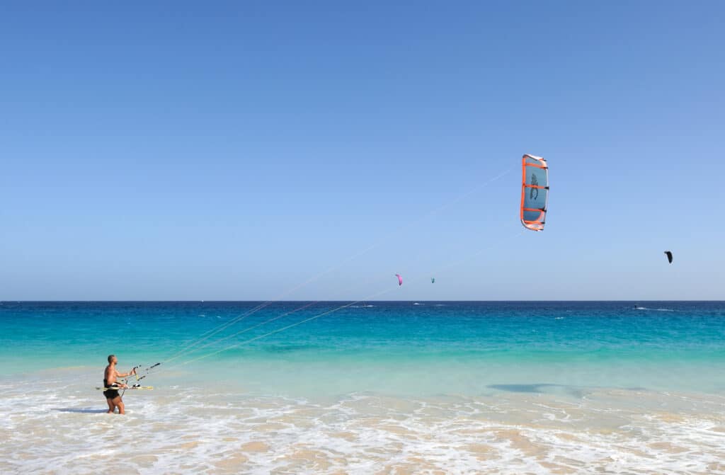 Homem na área rasa da praia praticando kitesurf. O mar tem poucas ondas e é cristalino, com um tom degrade azul