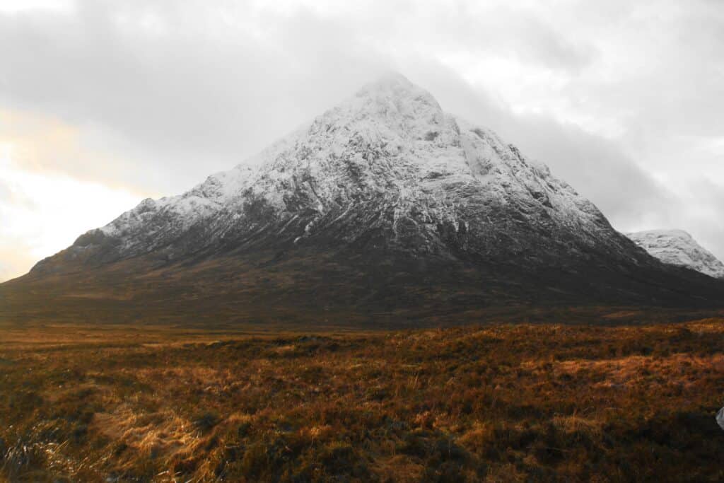 Um grande montanha das Terras Altas Escocesas com topo afunilado e cheio de neve. O céu está nublado e mais próxima da câmera é possível ver a grama outonal em tons marrom e vermelho