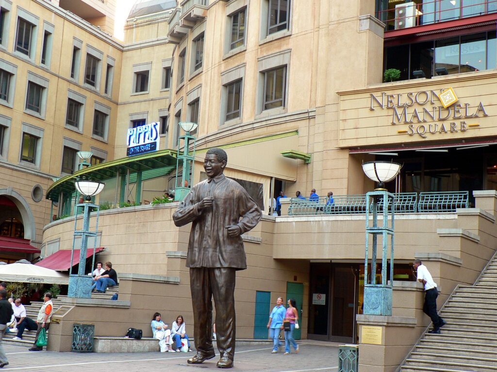 Estátua de Nelson Mandela dançando em frente ao shopping da Praça Nelson Mandela para ilustrar o post sobre chip de celular para Johanesburgo. Há pessoas caminhando ao redor e alguns prédios bege atrás da estátua. - Foto: misio via Pixabay