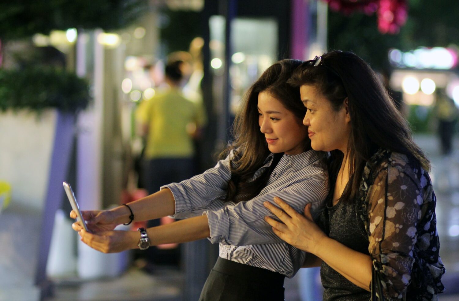 Duas mulheres de origem tailandesa tirando uma selfie juntas em uma das ruas de Bangkok, uma delas é mais velha e a outra é uma adolescente e é quem está segurando o celular para a foto