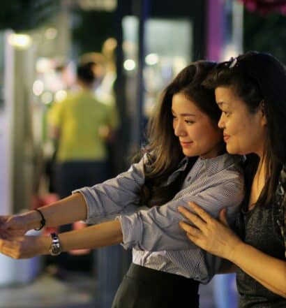Duas mulheres de origem tailandesa tirando uma selfie juntas em uma das ruas de Bangkok, uma delas é mais velha e a outra é uma adolescente e é quem está segurando o celular para a foto