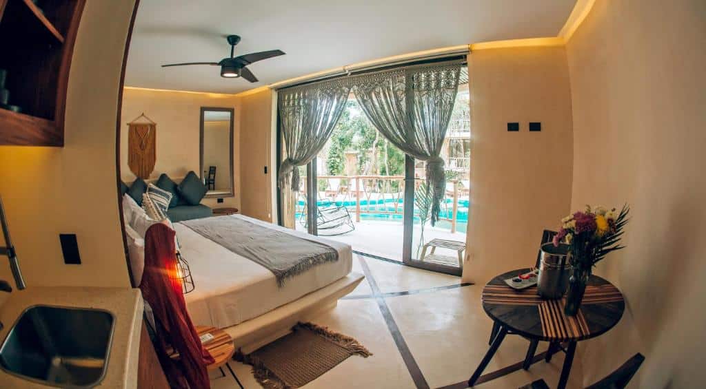 Suíte do Kan Tulum, de 39 m², com cama de casal, sofá no lado esquerdo, mesinha redonda com vaso de flor e balde de alumínio no lado esquerdo. À frente há uma porta de vidro com com cortina artesanal e vista para a piscina