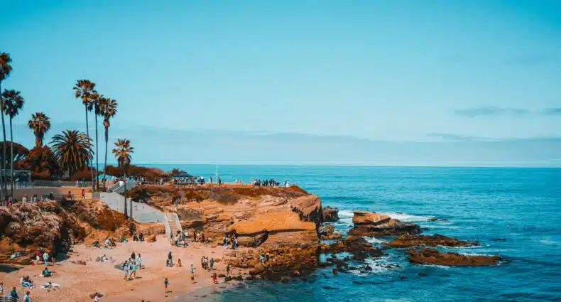 Praia de La Jolla, em San Diego. Na imagem é possível ver pedras do lado direito, cheias de pessoas, alguns bancos e escadas. Logo em frente, do lado esquerdo, está o mar. A foto ilustra o post de pontos turísticos em San Diego.