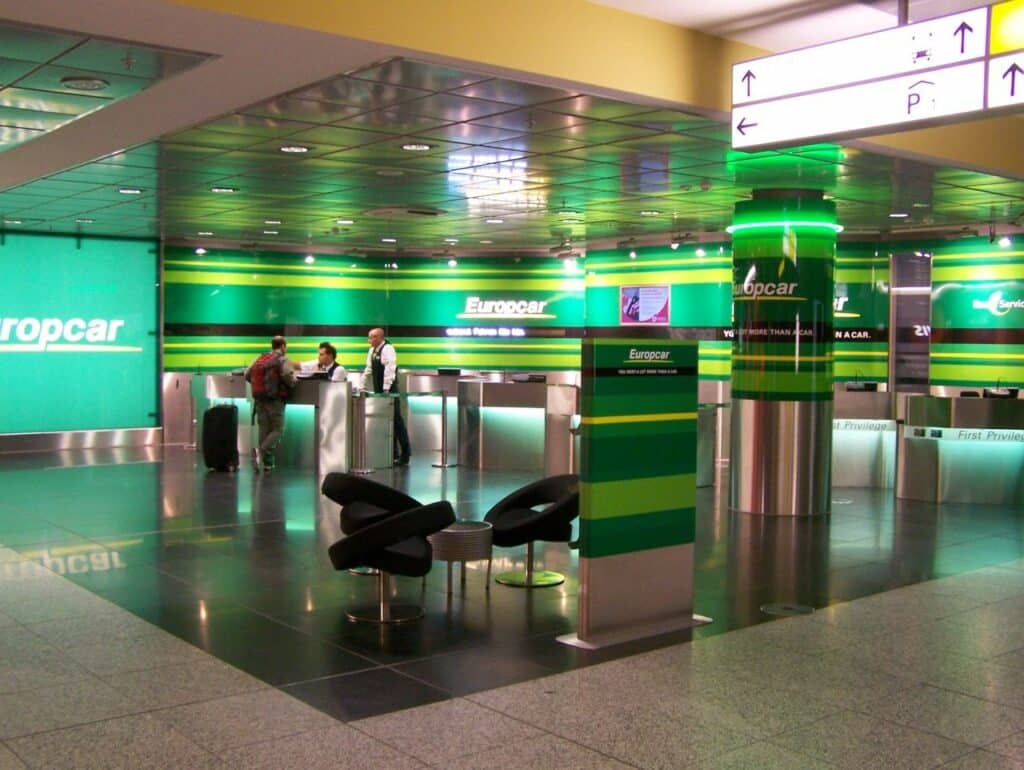 Loja da Europcar com decoração toda em verde, incluindo as paredes, e balcões metálicos, sendo que em um deles há uma pessoa com mochila e mala de rodinhas sendo atendida por dois atendentes da marca
