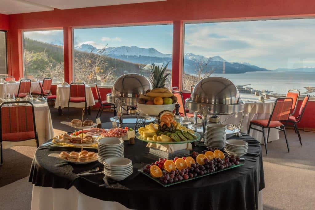 refeitório do Los Acebos Ushuaia Hotel com mesa farta de alimentos e parede de vidro com vista para as montanhas e o lago
