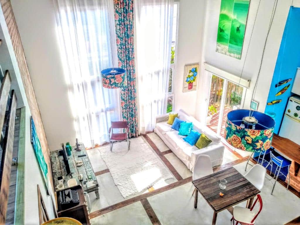 Sala de estar ampla na Maison de la Mer Espaço Inteiro praia 250m com um sofá com almofadas coloridas, janelas com cortinas brancas e coloridas, uma mesa de madeira com três cadeiras, televisão e alguns itens de decoração, para representar pousadas na Praia do Campeche