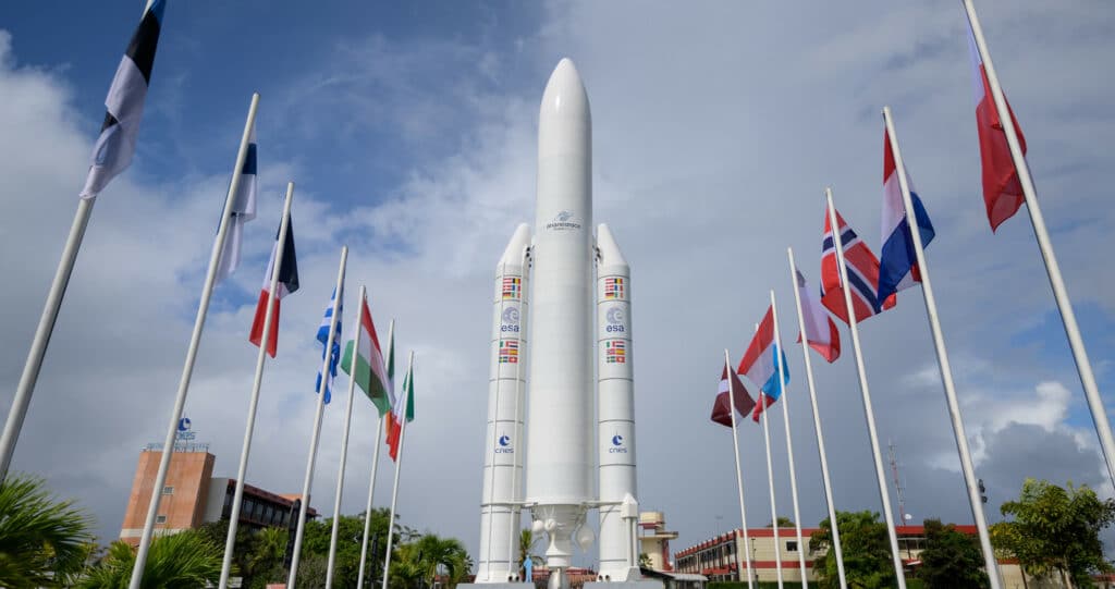 Maquete de um foguete, com bandeiras de vários países enfileiradas à frente. Há prédios e árvores ao fundo, e o céu está azul, ensolarado e com nuvens brancas