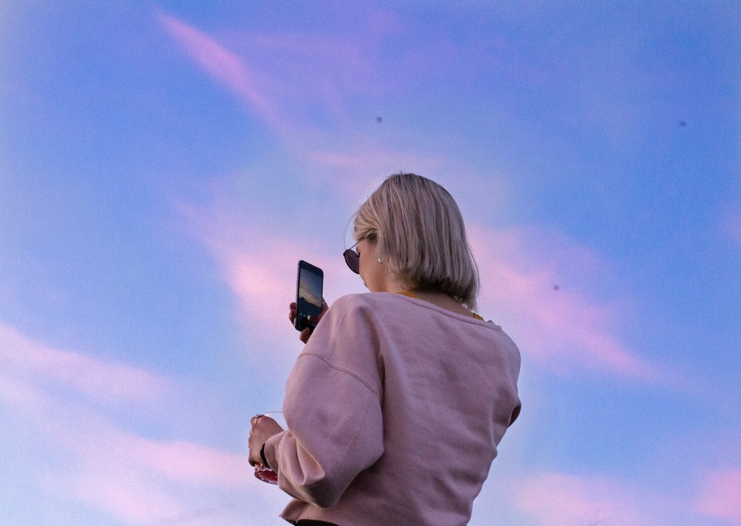 Mulher loira usando uma blusa de moletom cor de rosa enquanto segura o celular para tirar foto do céu azul e rosa. - Foto: Marie-Michèle Bouchard via Unsplash