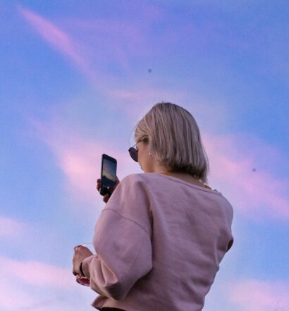Mulher loira usando uma blusa de moletom cor de rosa enquanto segura o celular para tirar foto do céu azul e rosa. - Foto: Marie-Michèle Bouchard via Unsplash