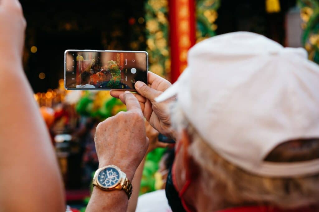 Um senhor com cabelos brancos segurando um celular na horizontal enquanto fotografa uma apresentação de rua em Bangkok