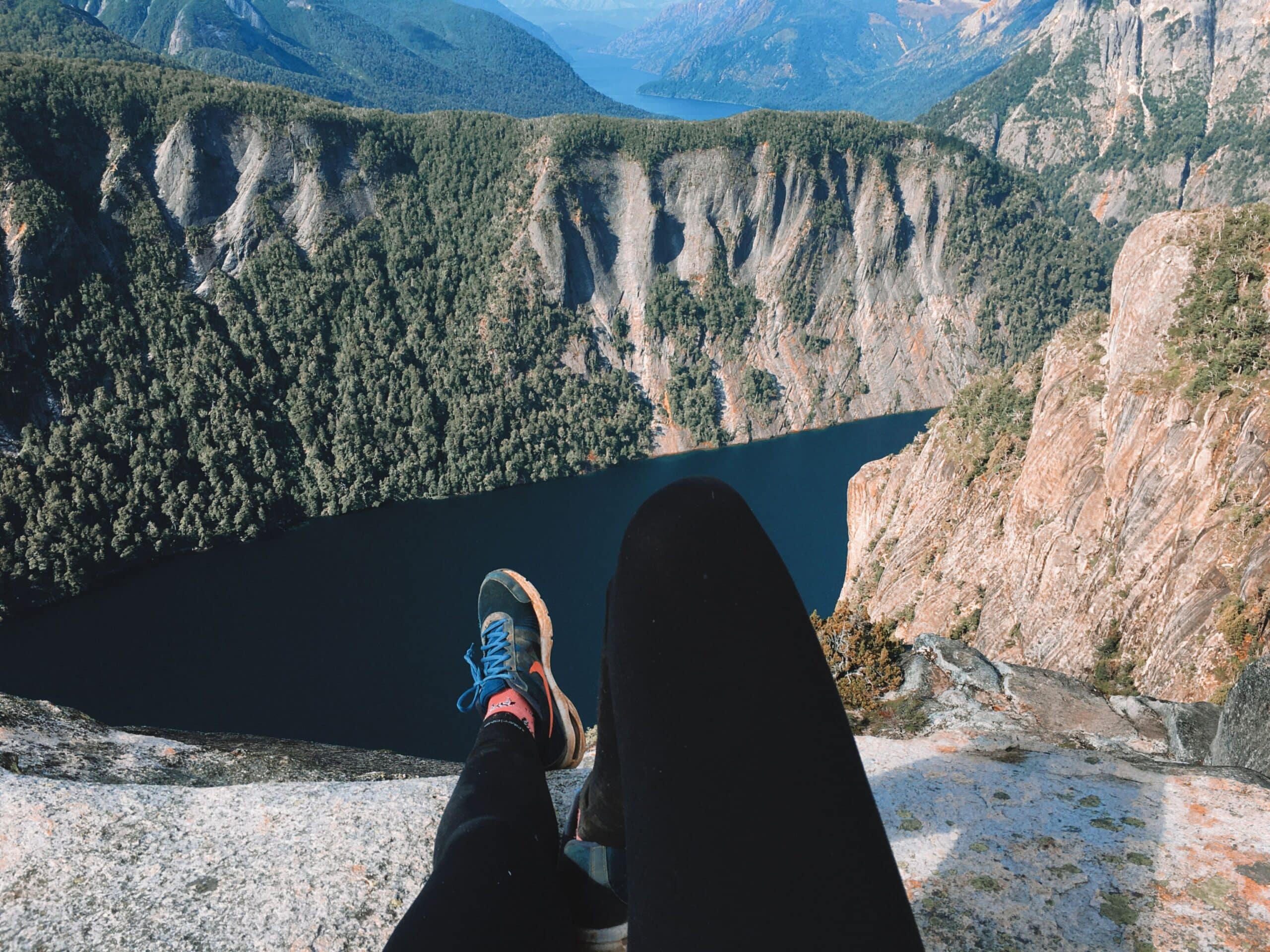 paisagem das montanhas em um vale de Bariloche com o rio Negro visto mais a frente. Próximo a câmera é possível enxergar duas pernas femininas com calça legging preta e tênis de trekking esticadas sobre a rocha