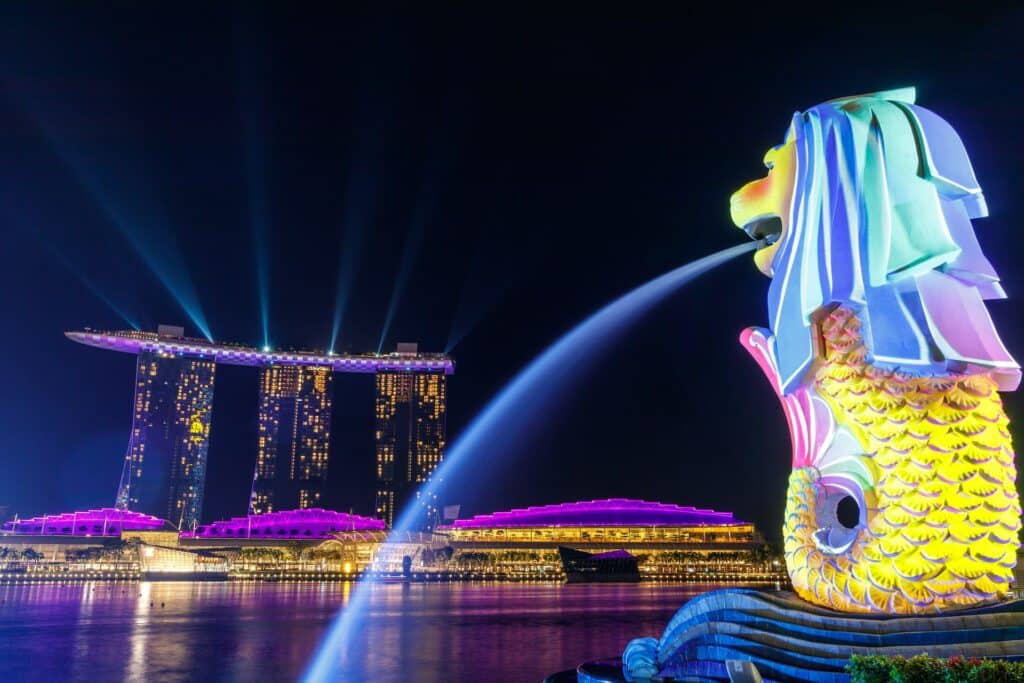 Noite iluminada, com a estátua "The Merlion", símbolo nacional com cabeça de leão e corpo de peixe, soltando um foco de luz em direção ao Rio Singapura e com vários prédios iluminadas ao fundo.