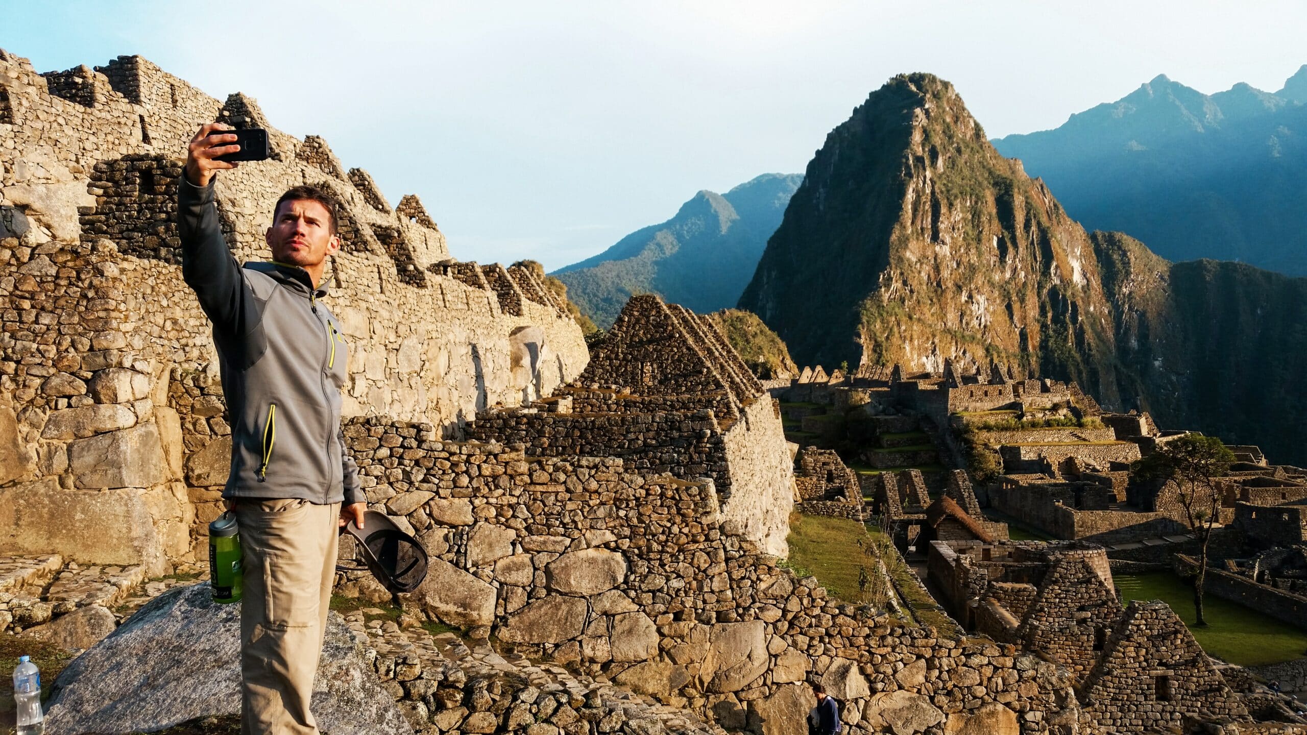 homem branco tirando uma selfie com seu celular em Machu Picchu, no Peru, com toda a paisagem natural do sítio arqueológico atrás de si