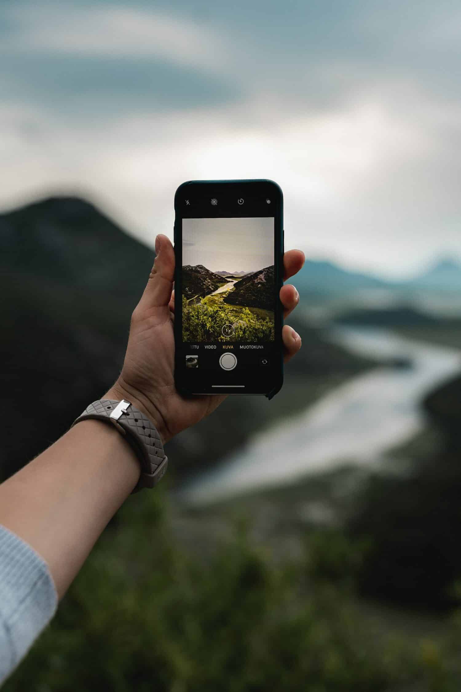 Uma mão branca com um relógio segurando um celular na vertical enquanto fotografa uma rio passando por entre duas montanhas