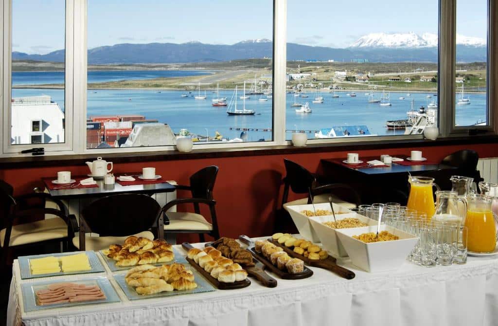 refeitório do MIL810 Ushuaia Hotel com vista para o lago e as montanhas, e uma mesa repleta de alimentos para café da manhã, como pães, frios e sucos